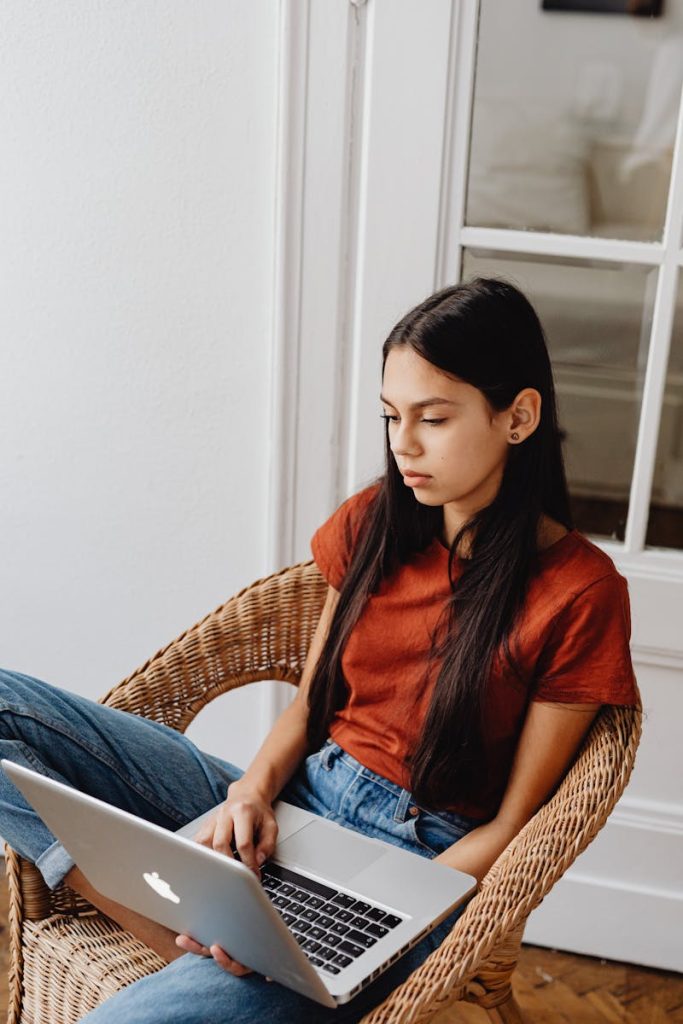 Young girl comfortably sitting indoors, focused on her laptop for casual browsing or online activity.
