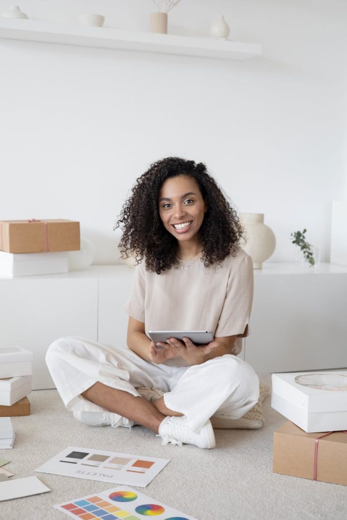 A confident young woman sitting among packages and papers while using a tablet, showcasing modern entrepreneurship.